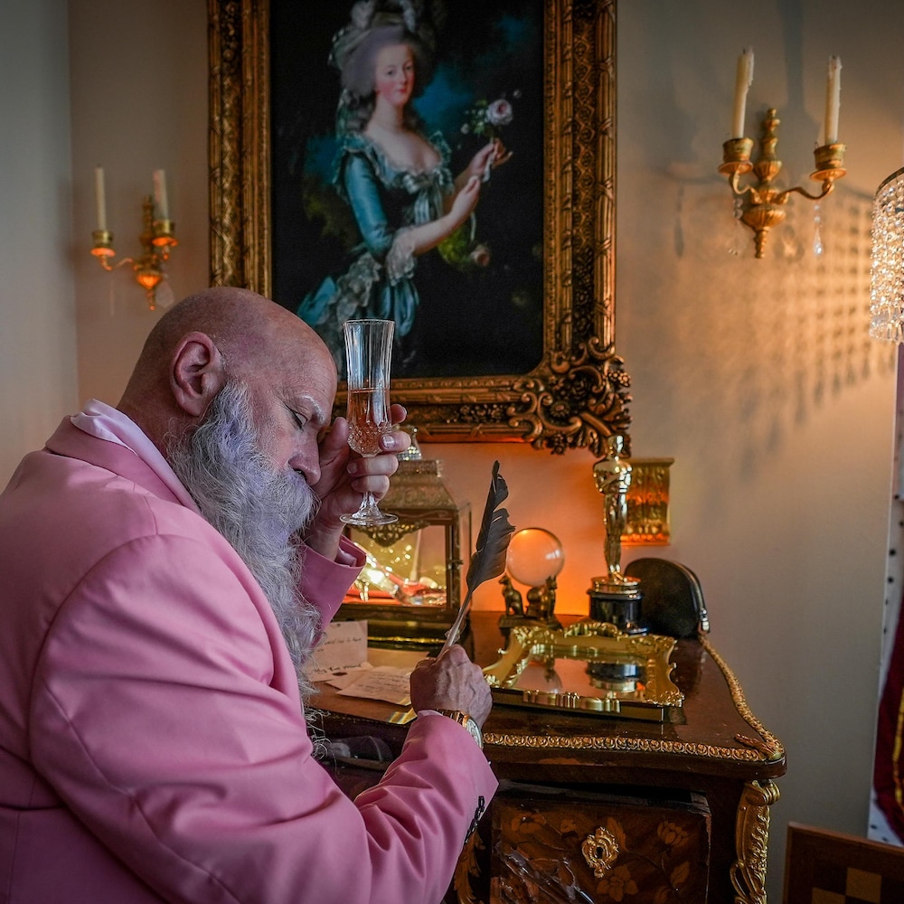 Mark Henderson assis à son bureau, une plume et une coupe de cristal à la main. Un portrait de Marie-Antoinette est encadré et exposé au mur. Un oscar et une pantoufle de verre sont déposés sur son bureau.