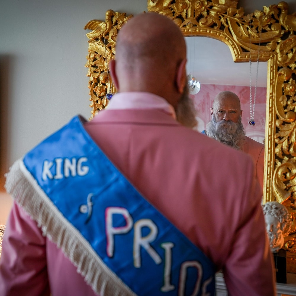 Mark Henderson se regarde dans un miroir. Il porte un costume rose et une bannière autour du tronc qui dit : Roi de la Fierté.
