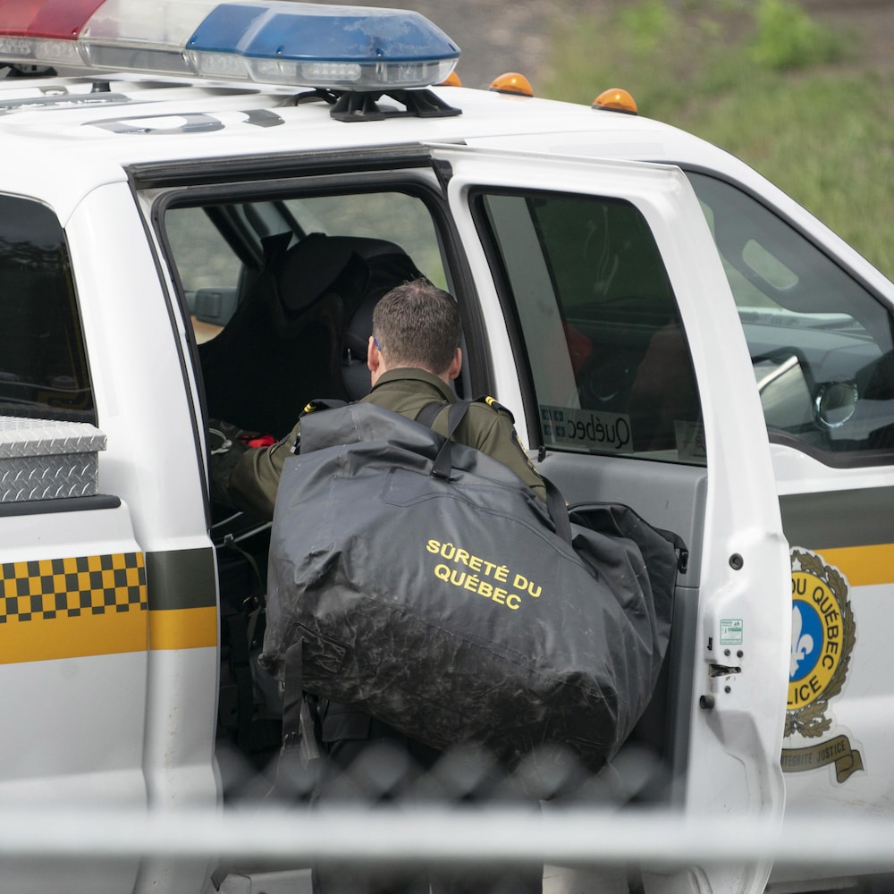 Un policier, portant un gros sac sur son épaule, fouille dans la banquette arrière d'un véhicule de la Sûreté du Québec. 