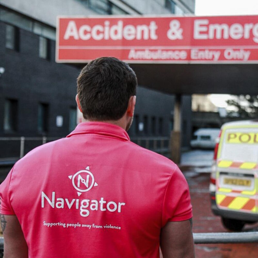 Un homme de dos, arborant un chandail rose du programme des Navigateurs. Il est à l'extérieur, devant l'urgence d'un hôpital.