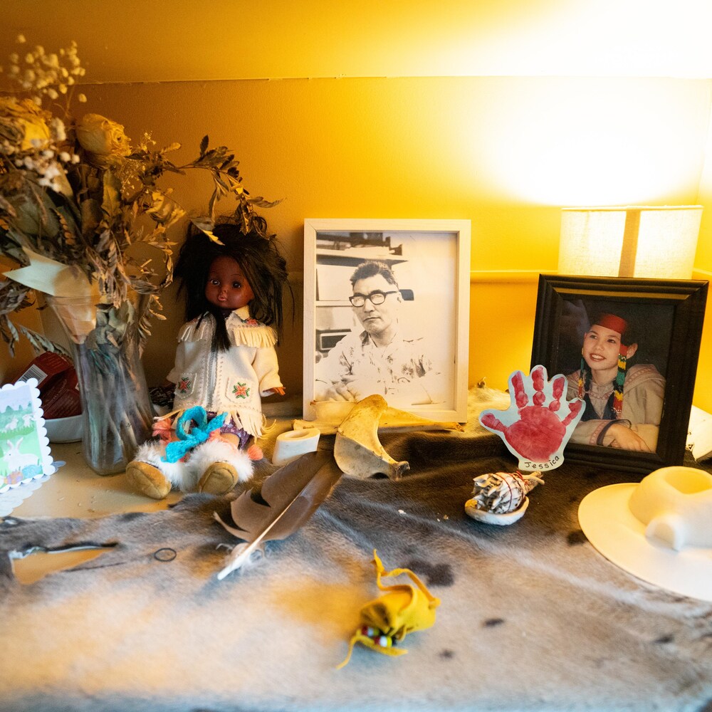 Des objets sur une table : un bouquet de fleurs séchées, des cadres avec photos, une poupée, des cartes, un chapeau, une plume, etc.