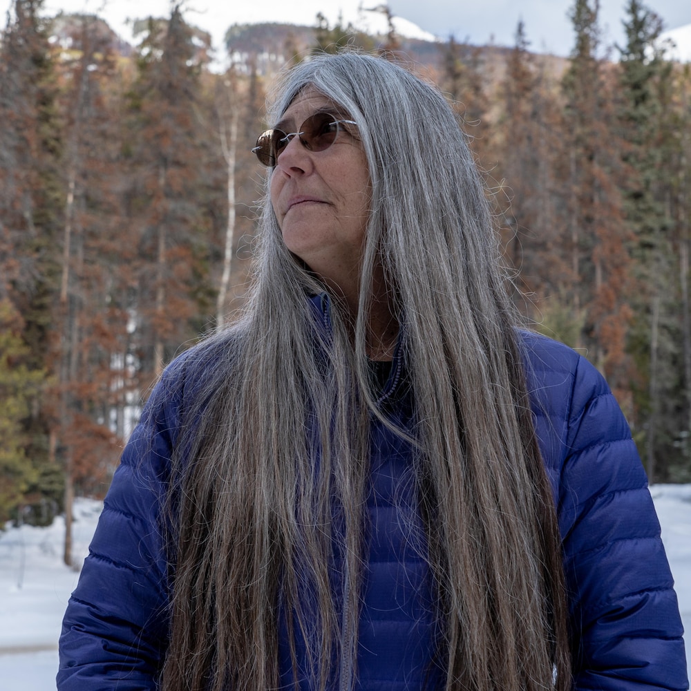 Une femme dans la soixantaine aux cheveux longs gris et avec des lunettes fumées pose de profil devant une forêt de conifères. 