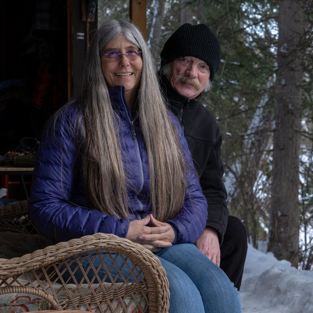 Une femme et un homme dans la soixantaine souriants pose assis sur un banc en rotin. Une forêt est à l'arrière-plan.