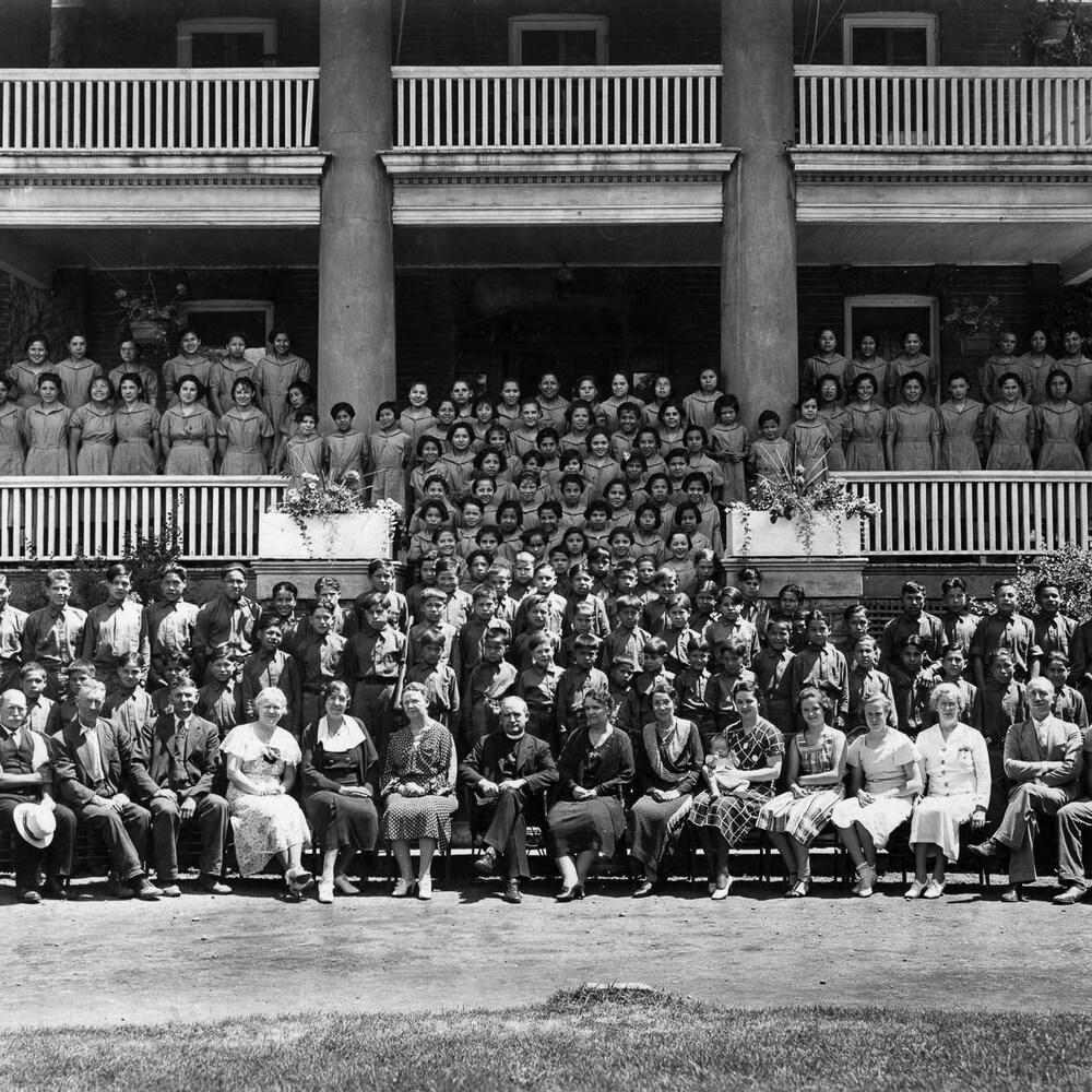 Une foule de jeunes des Premières Nations se tiennent les bras croisées dans le dos pour la photo, leur professeurs blancs assis à l'avant