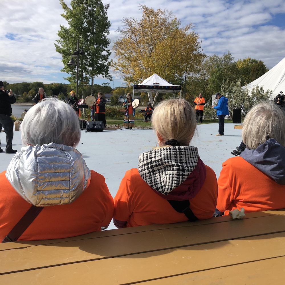 Trois dames portant des gilets de couleur orange écoutent un spectacle.
