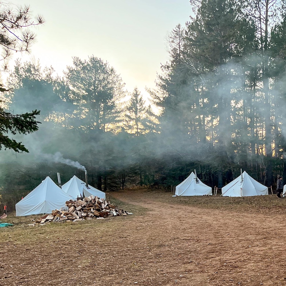 Huit tentes blanches avec des cheminées dressées près d’un boisé.