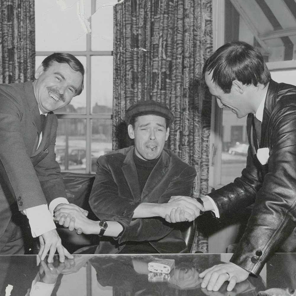 Jacques Brel assis, serrant les mains d’Albert Larouche et de Jacques Houde.