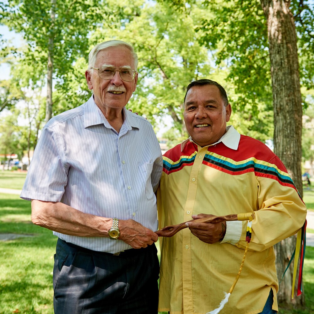 Raymond Currie et un homme en habits autochtones colorés se trouvent dans un parc et tiennent un bâton de parole.