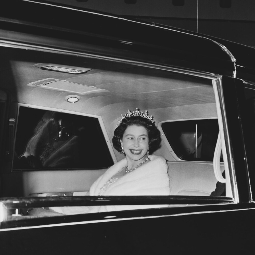 Une jeune reine Élisabeth II sourit en regardant par la vitre arrière d'une grande voiture noire.