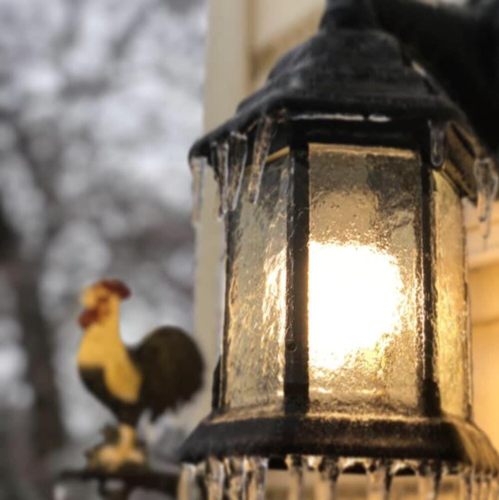 Une lumière extérieure, allumée, recouverte de verglas. Une figurine de coq floue est en arrière-plan.