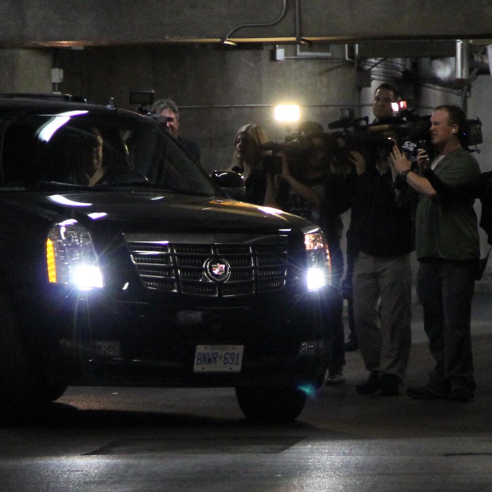 Le maire, au volant de son véhicule, entre dans le stationnement souterrain devant une foule de journalistes qui l'attendent.