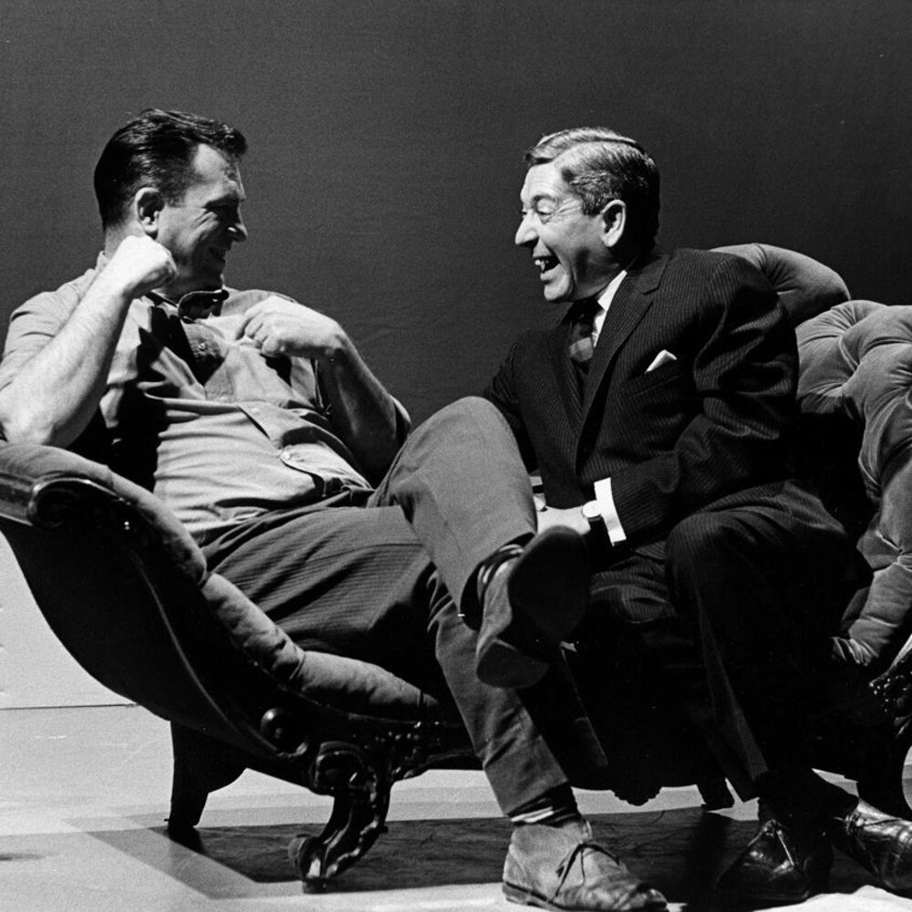Dans un studio de télévision, l'écrivain américain Jack Kerouac et l'animateur Fernand Seguin, riant, sont assis sur une causeuse.