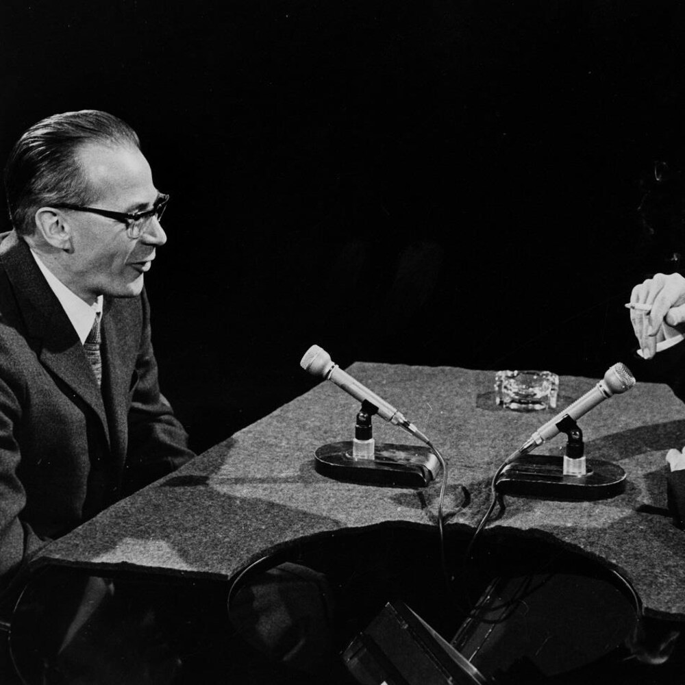Dans un studio de télévision, Léon Dion s'entretient avec l'animateur Fernand Seguin. Deux microphones sont posés sur le pupitre devant eux.
