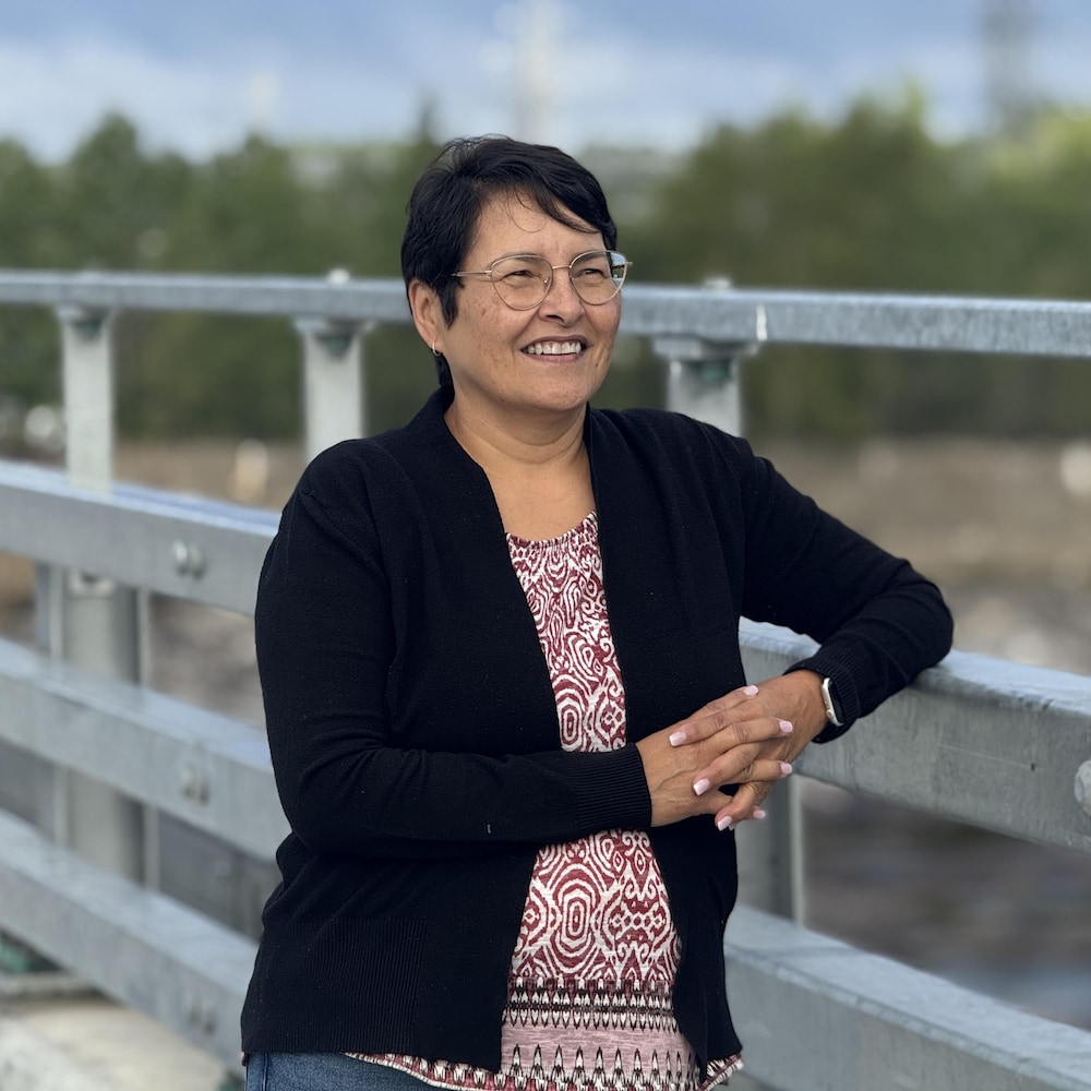 Une femme souriante prend appui sur la rambarde métallique d’un pont, devant un paysage boisé.