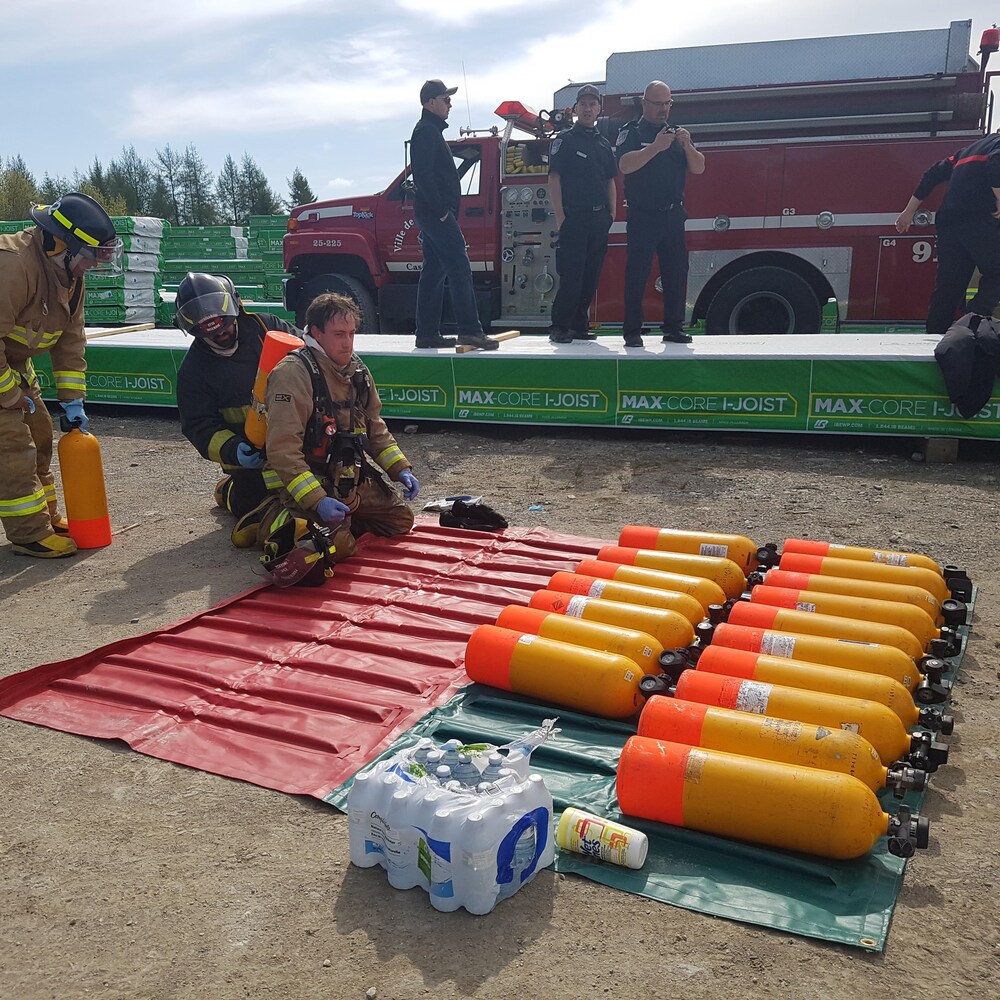 Un pompier est à genoux alors que son collègue lui installe une bonbonne d'oxygène dans le dos.