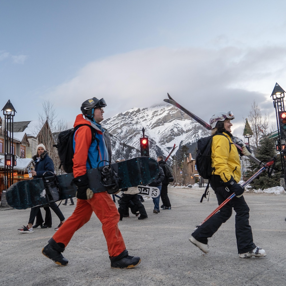Un couple formé d'un skieur et d'une planchiste traverse une rue et on voit à l'arrière plan une énorme montagne enneigée. 