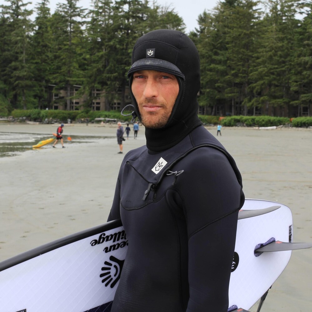 Peter DeVries en combinaison tient sa planche sous le bras, sur une plage.
