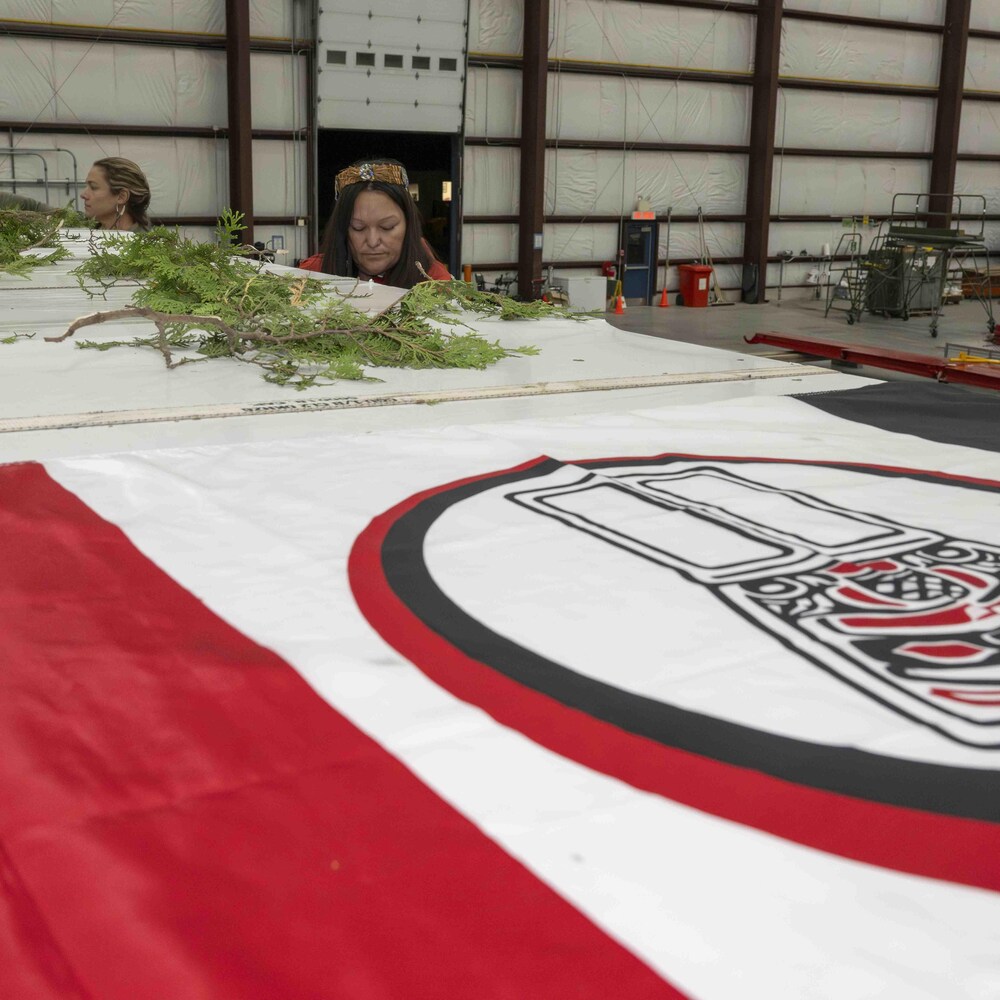 En avant-plan un drapeau de la nation Nisga'a et en arrière-plan deux femmes mettent du cèdre sur un caisson blanc
