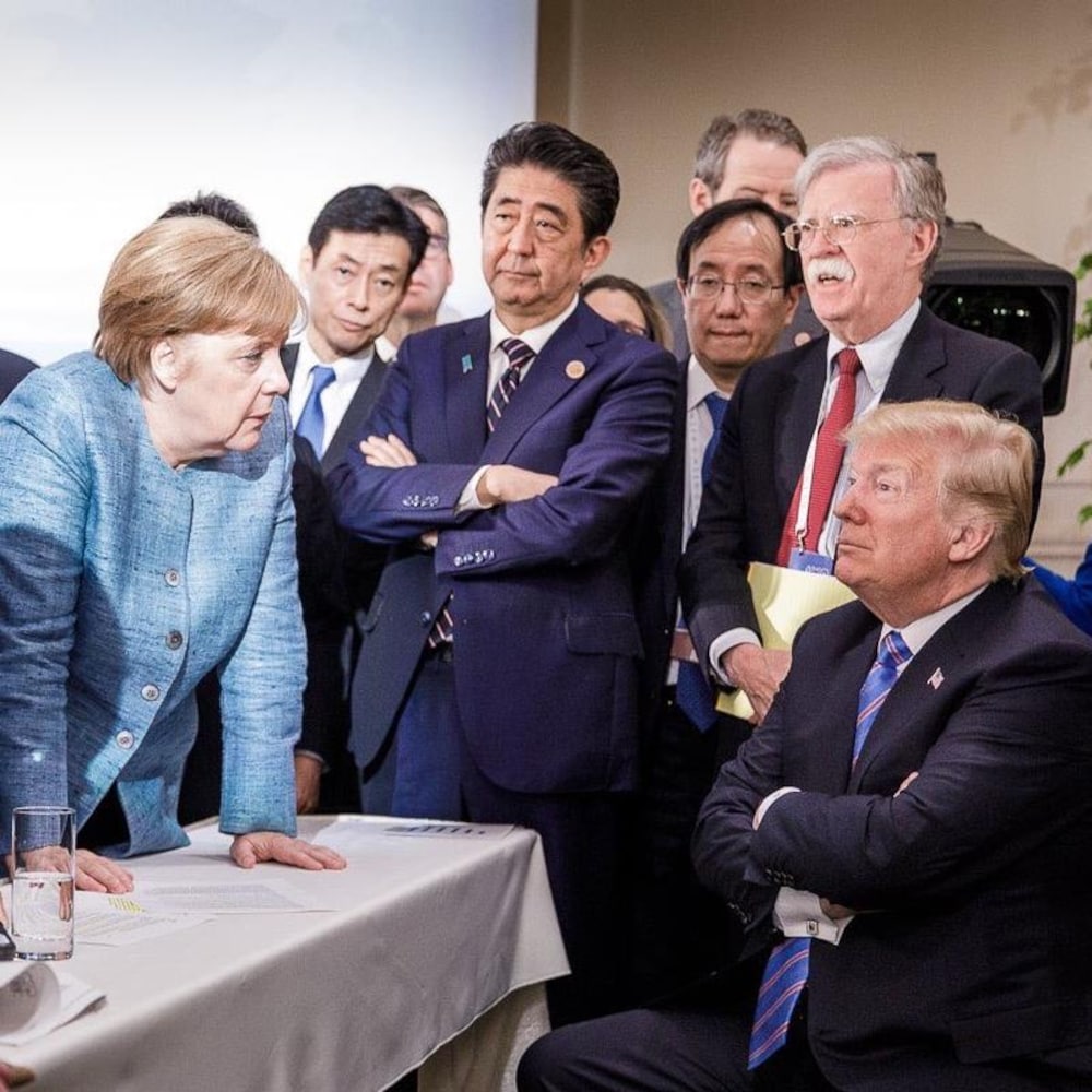 Donald Trump, assis, semble se faire gronder par Angela Merkel, debout devant lui. 