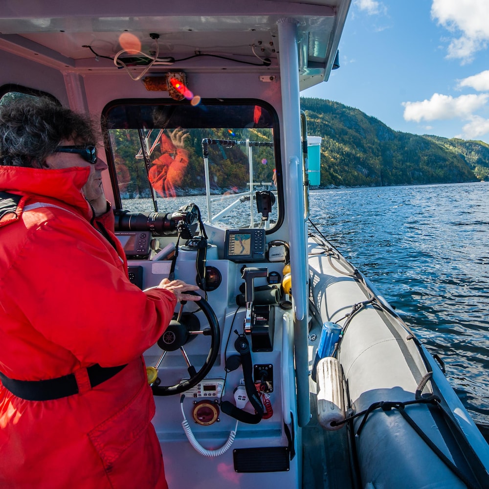 Robert Michaud, vêtu d'un manteau rouge, est à la barre du bateau pneumatique. On aperçoit, en arrière-plan, les montagnes du fjord. 