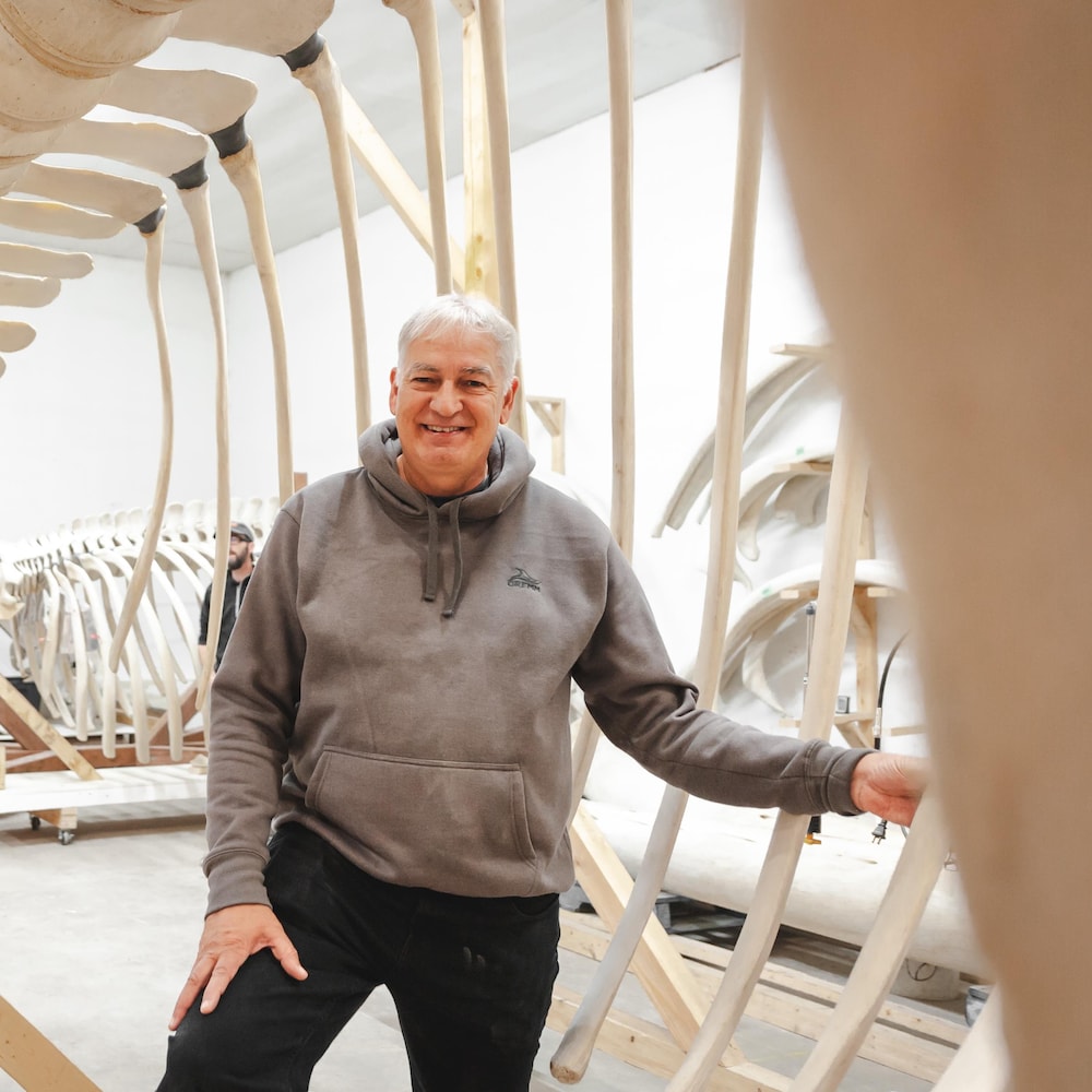 Patrice Corbeil, un homme dans la cinquantaine aux cheveux blancs, pose, souraint, à l'intérieur d'un squelette de baleines.