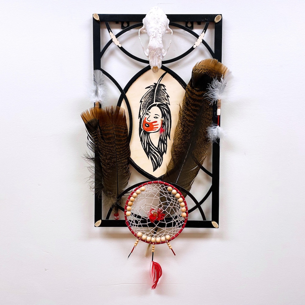Un cadre avec dedans des plumes, un attrape-rêves et un dessin d'une femme avec une main rouge sur la bouche.