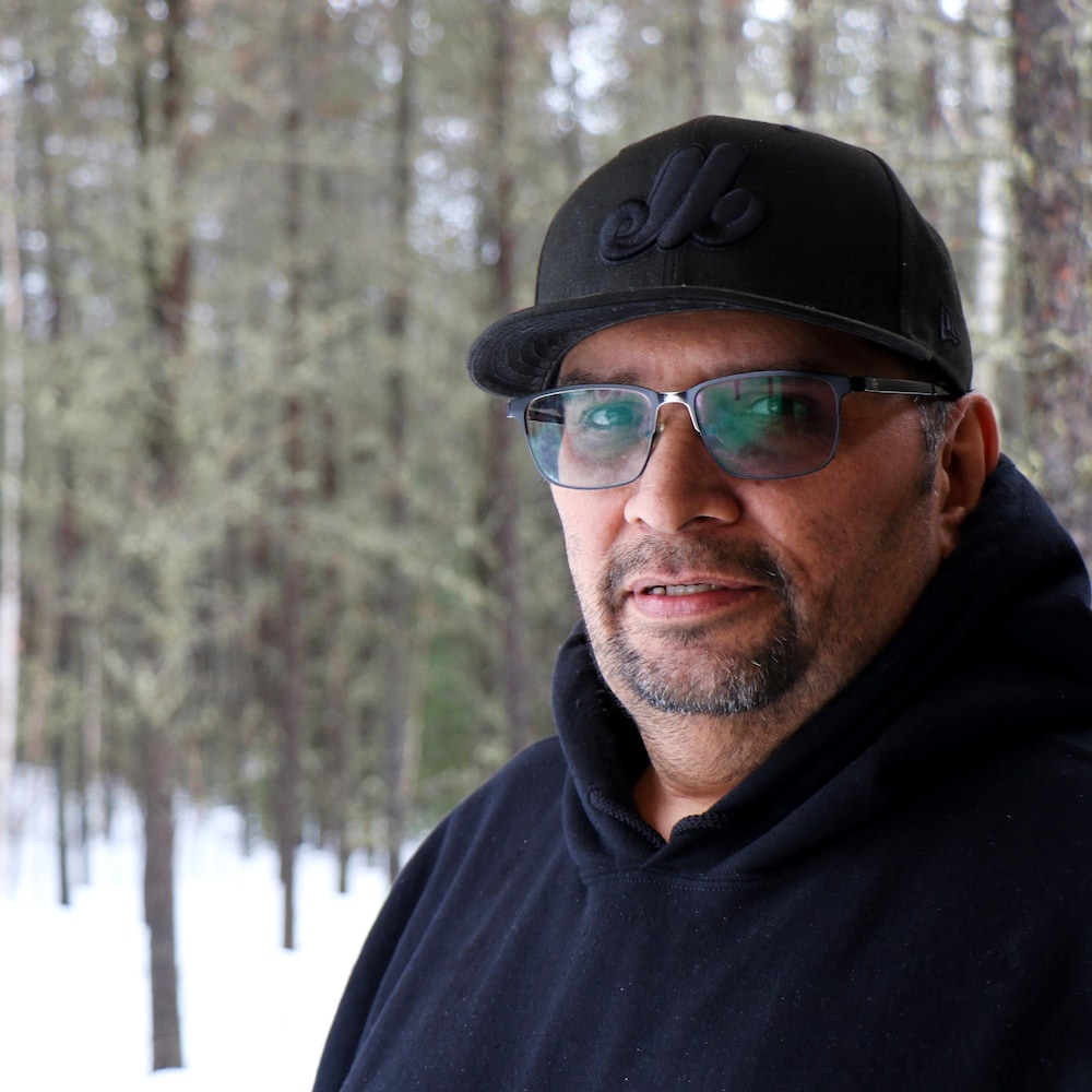 Un homme de face portant une casquette noire et un coton ouaté noir, dans la nature (bois et neige derrière lui).
