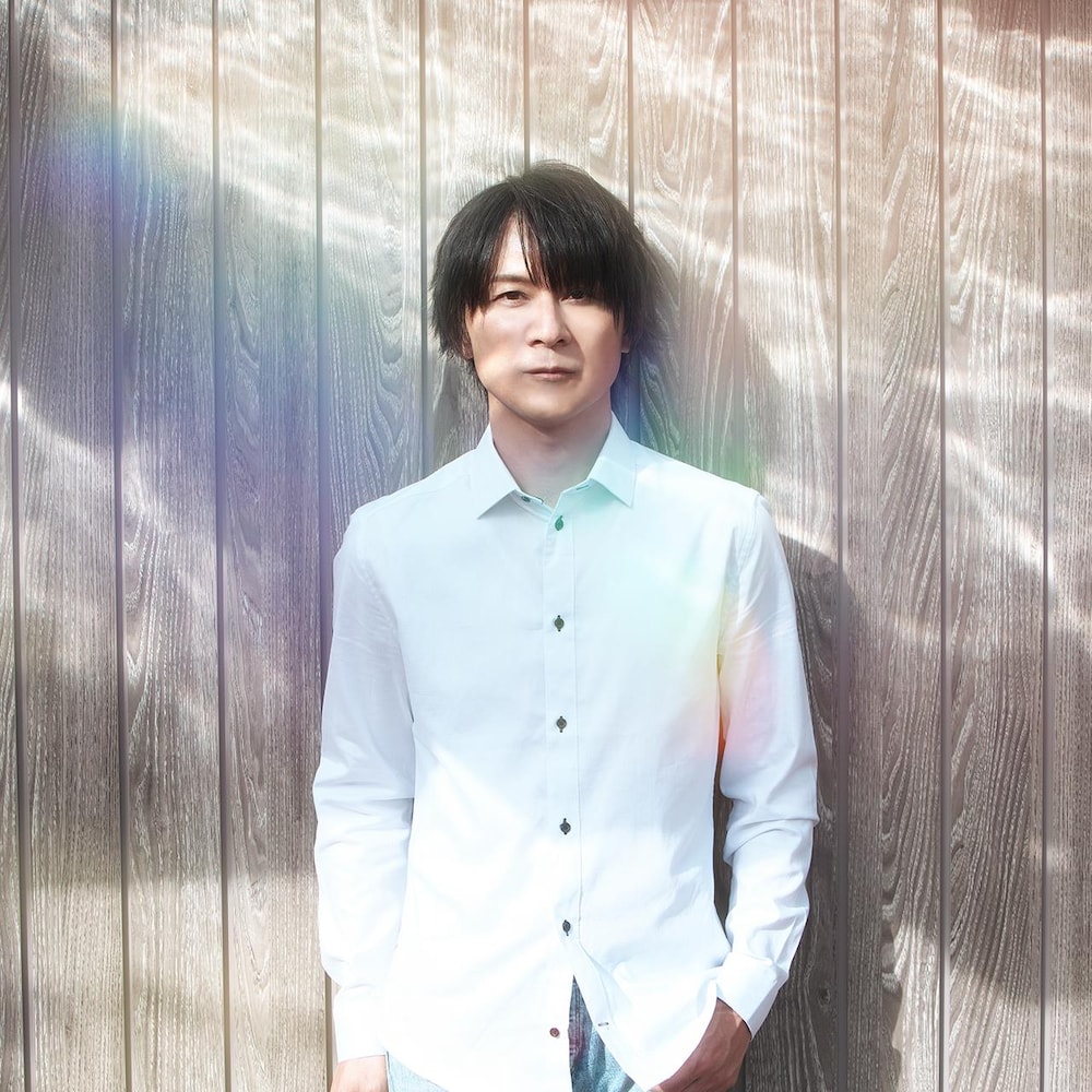 Yasunori Mitsuda est un homme dans la trentaine qui pose pour la caméra. Il porte une chemise blanche et ses longs cheveux foncés tombent au-dessus de ses yeux.