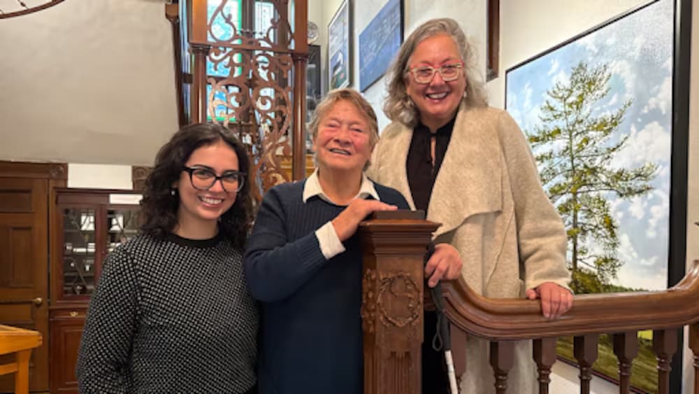 Trois femmes de trois générations différentes sourient ensemble, posant près de la rampe en bois sculpté d'un escalier intérieur, dans un cadre chaleureux et artistique de la Galerie 78, avec un tableau visible en arrière-plan.