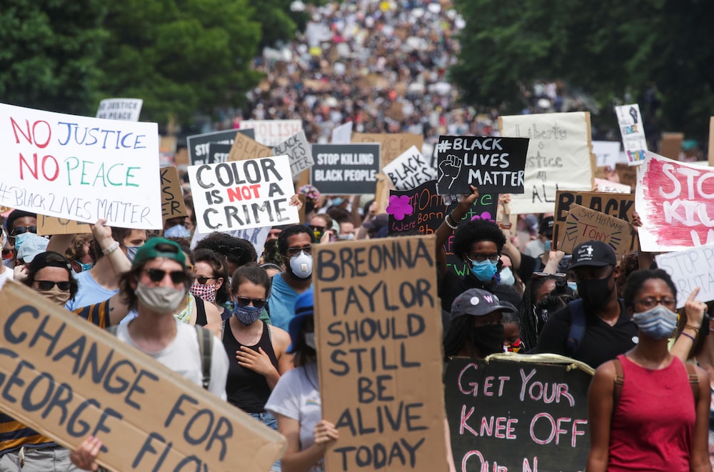 Une marée bigarrée de manifestants portant le masque et des pancartes occupent toute l'image.