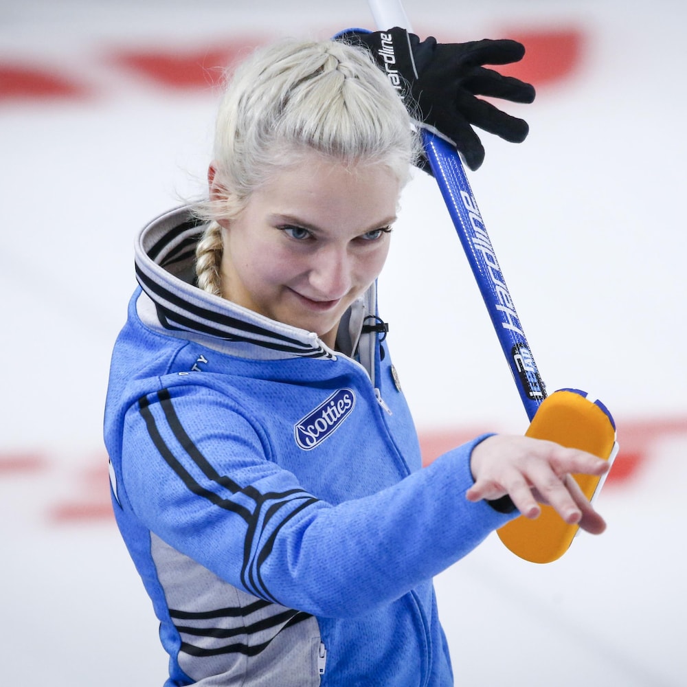 Une joueuse de curling tient son balais à la hauteur de sa tête, en bougeant les doigts. 