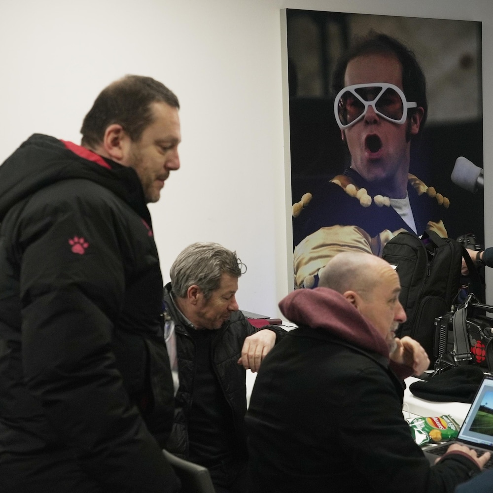 Des journalistes devant leurs ordinateurs dans une salle de presse au fond de laquelle on voit une affiche d'Elton John en spectacle