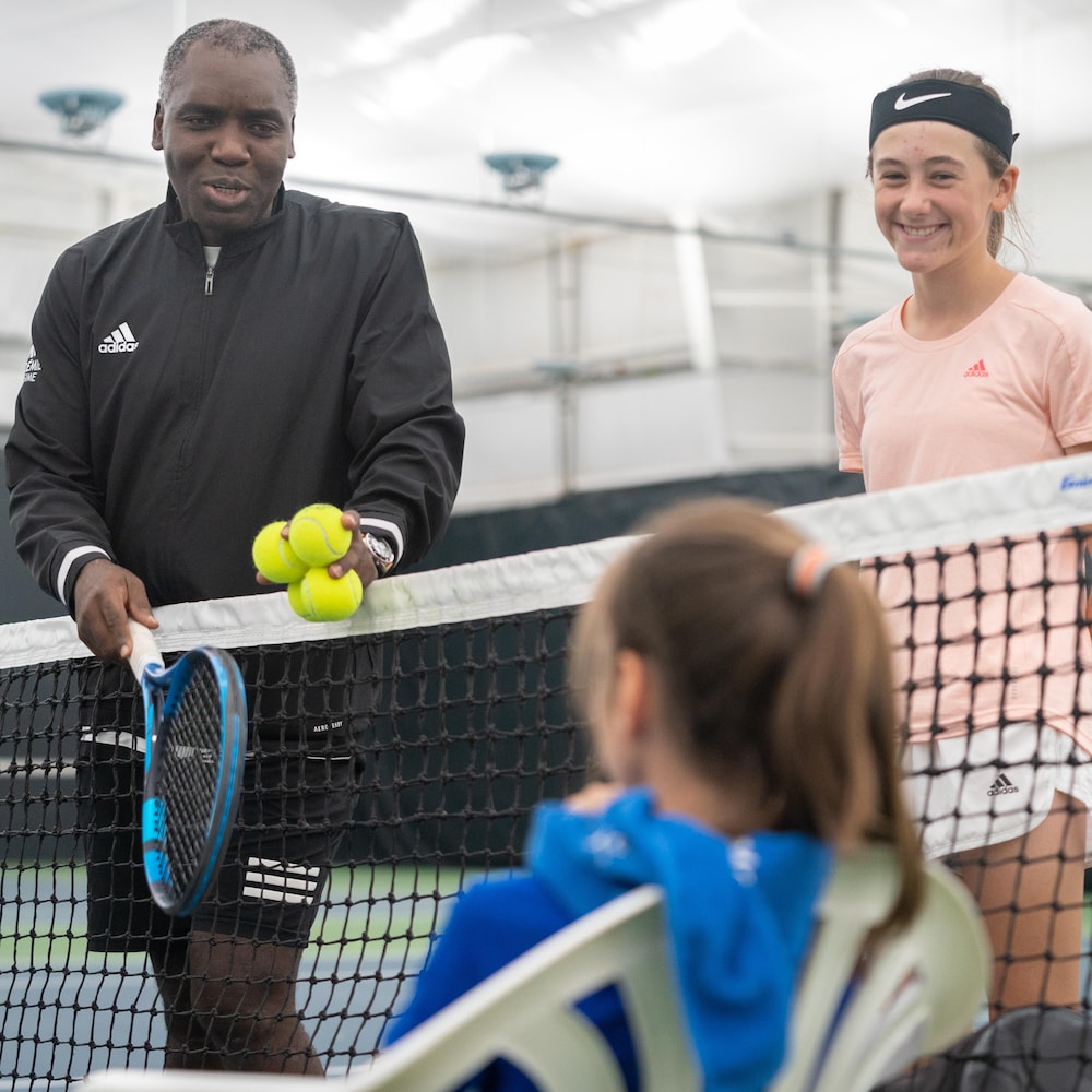 Un homme avec une raquette et des balles de tennis se tient près du filet pour discuter avec deux jeunes joueuses