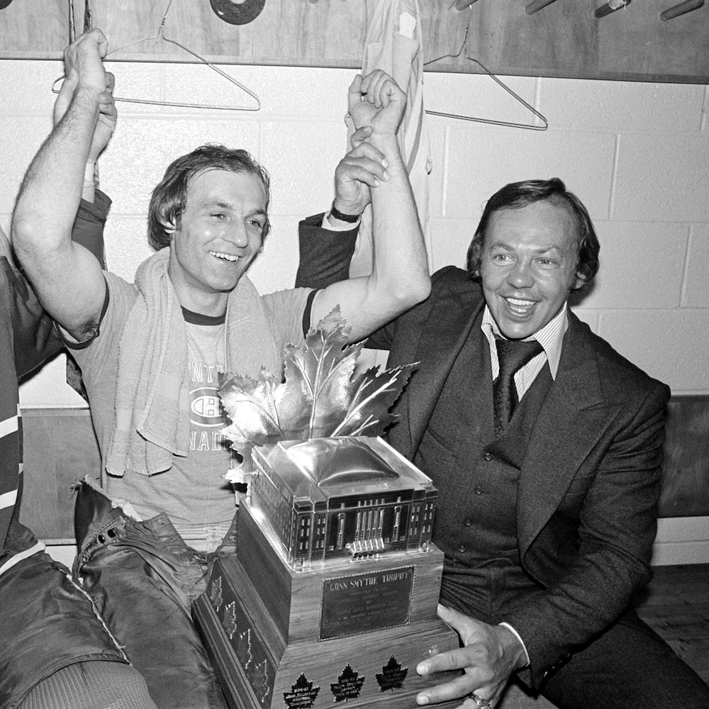 Un joueur de hockey avec un trophée sur ses genoux est félicité par deux coéquipiers dans le vestiaire de son équipe. 