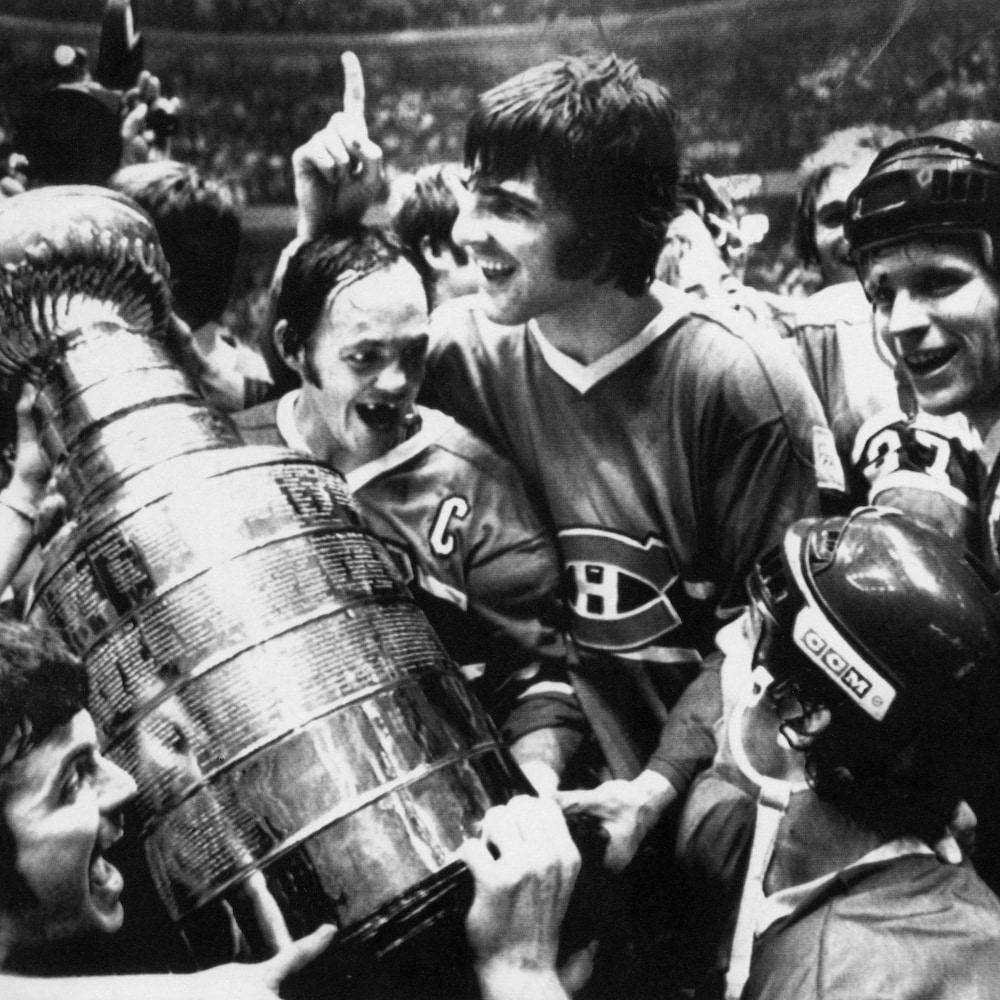 Photo en noir et blanc d'un joueur de hockey qui sourit entouré de ses coéquipiers avec un trophée dans ses mains.