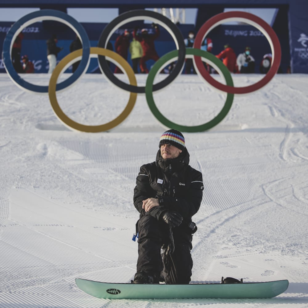 Un homme se tient un genou par terre sur une piste de surf des neiges, devant les anneaux olympiques.