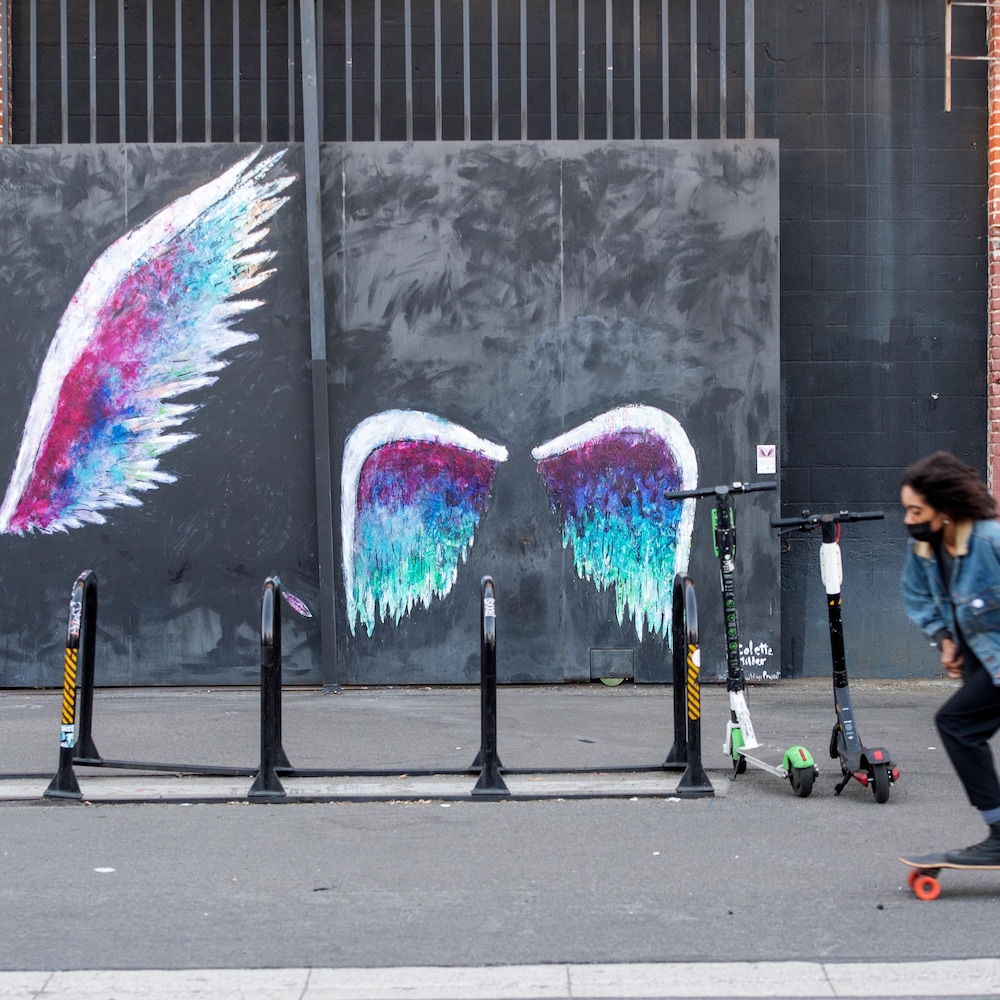 Quelqu’un portant un masque roule dans une rue déserte devant des graffitis représentant des ailes d’ange. 
