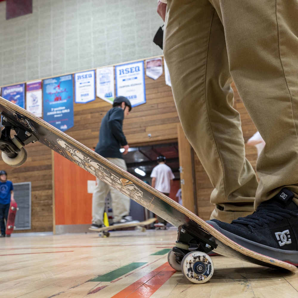 Dans un cours au gymnase, un élève maintient du pied sa planche en position inclinée, avec les deux roues du devant surélevées. 