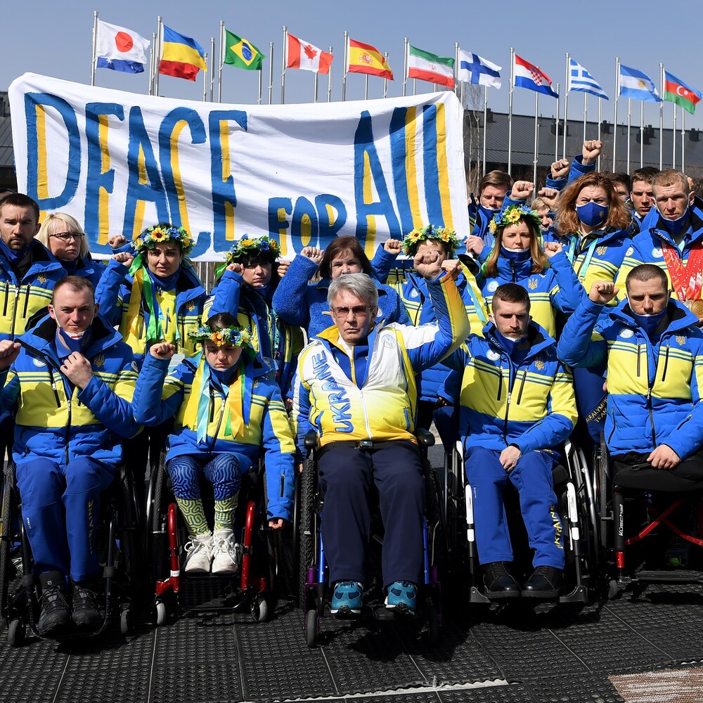 Un groupe de personnes dans des uniformes aux couleurs de l'Ukraine lèvent le poing droit et tiennent une affiche : « La paix pour tous » (peace for all). 