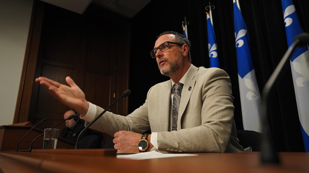 Jean-François Roberge tend son bras en point de presse devant des drapeaux québécois.
