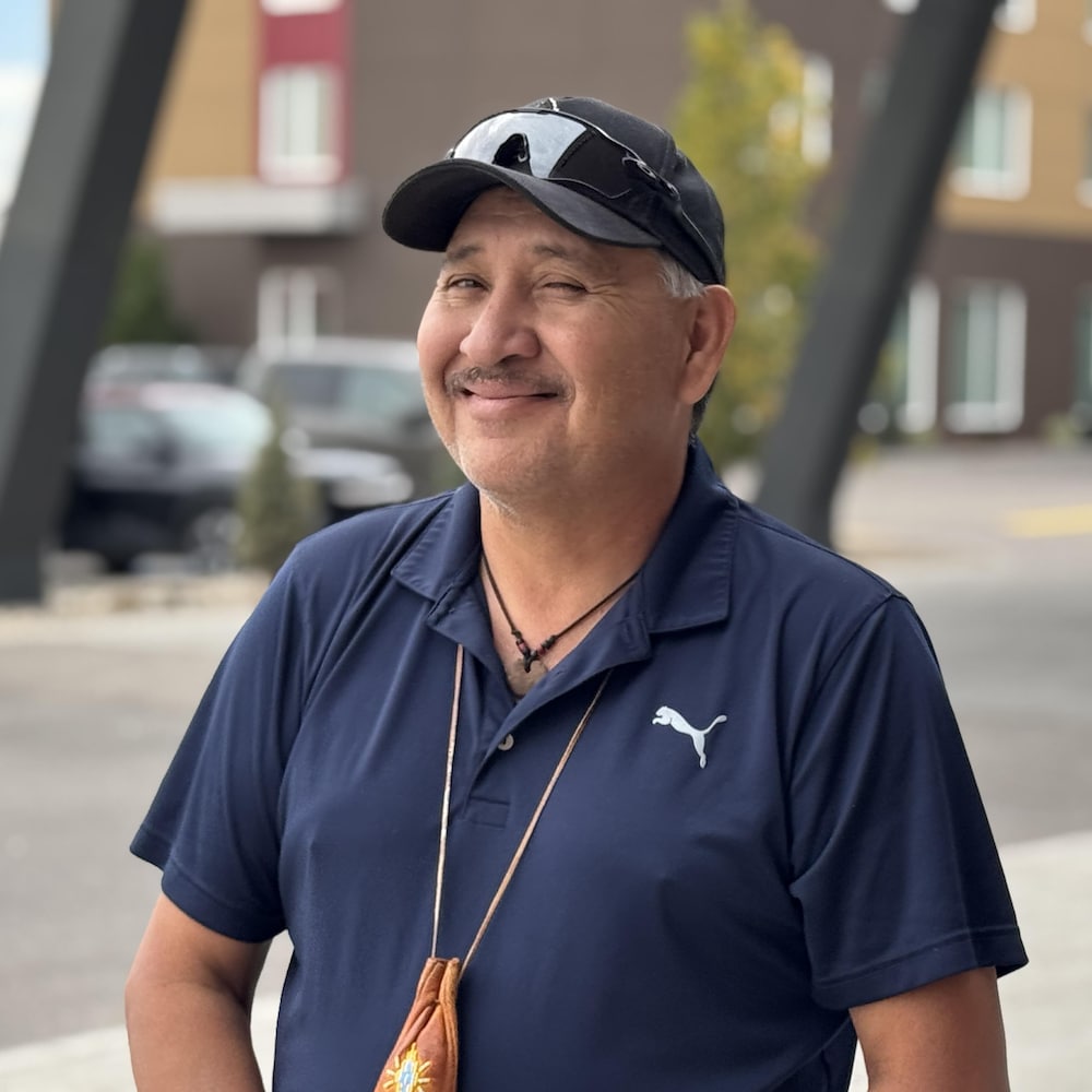 Un homme en polo bleu marine sourit, des lunettes de soleil posées sur sa casquette.