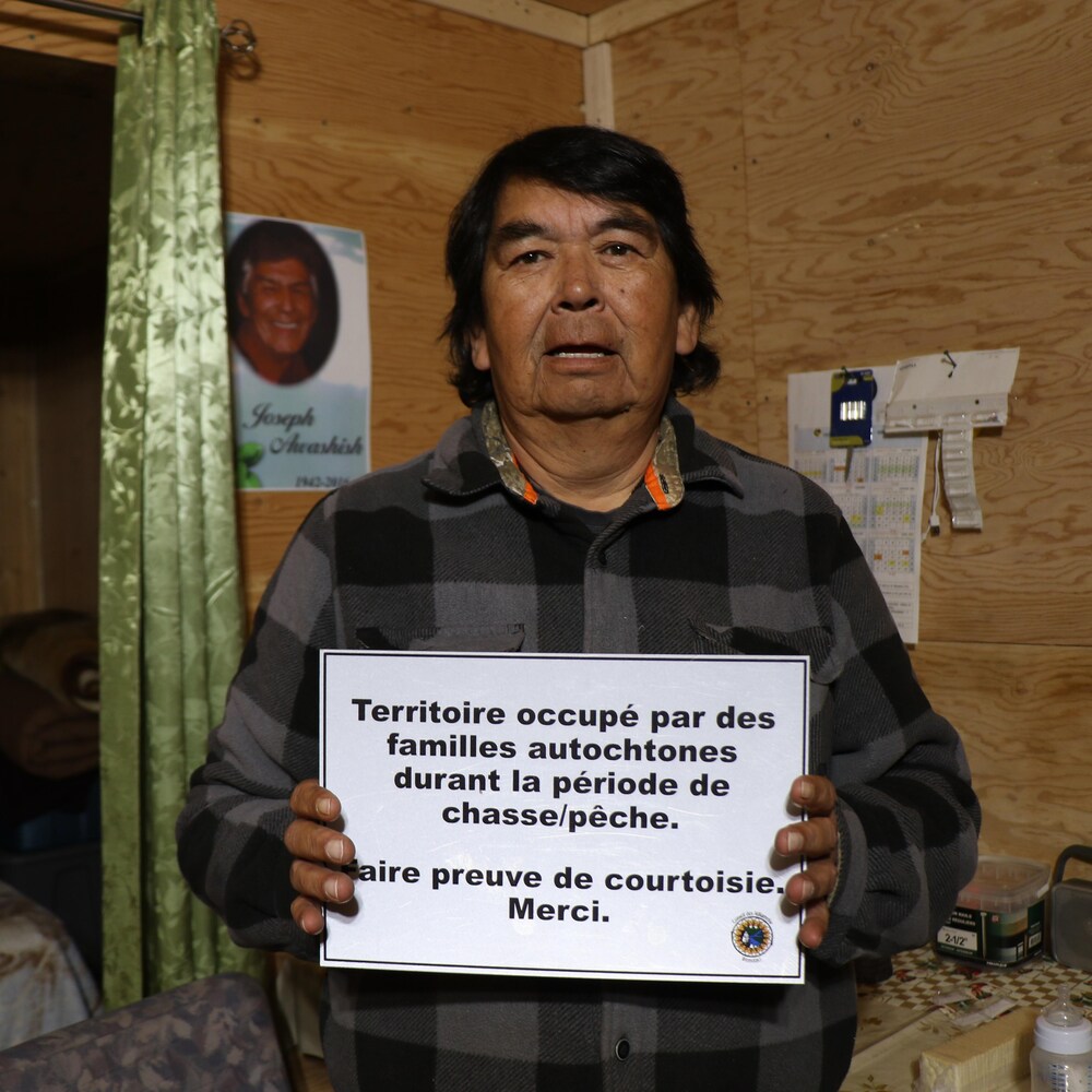 Un homme tient un panneau sur lequel est écrit : Territoire occupé par des familles autochtones durant la période de chasse-pêche, faire preuve de courtoisie. Merci.