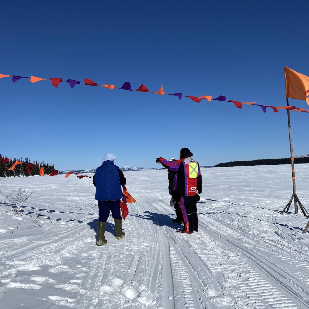 Trois personnes de dos sur un lac avec des montagnes au loin et au-dessus d'eux des drapeaux rouges, orange et mauves.