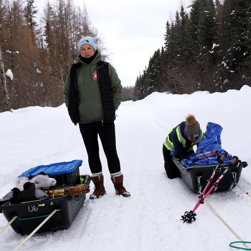 Une femme debout, entre deux traîneaux sur la neige, et une autre femme qui est à genoux à chercher quelque chose dans son traîneau. 