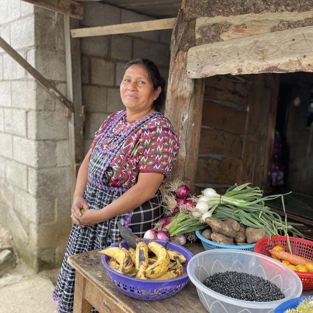 Rosa se tient devant un kiosque de fruits et de légumes frais, parmi lesquels on voit entre autres des carottes et des bananes. 