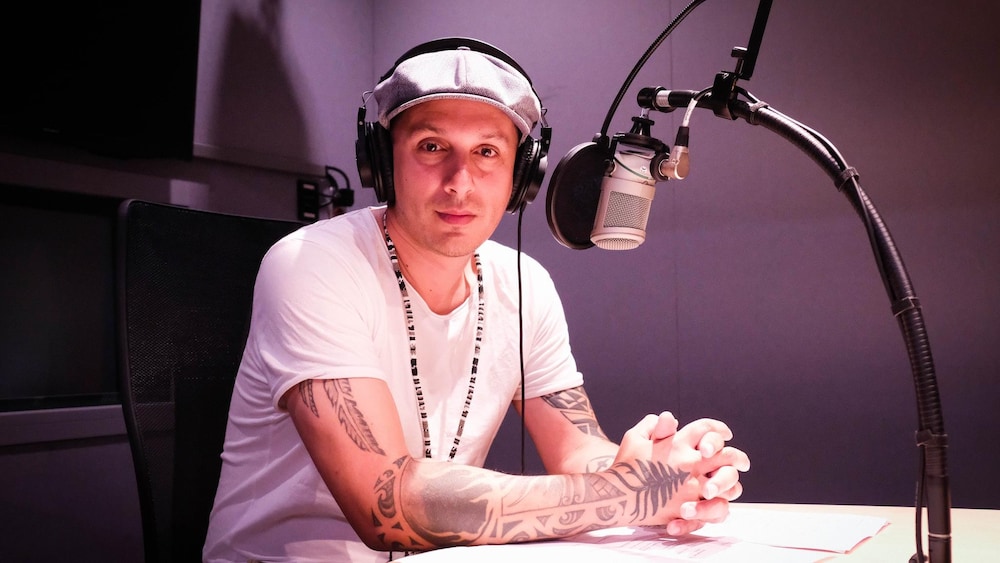 Samian pose devant un micro en studio. L'artiste est l'animateur de l'émission Minotan sur ICI Musique.