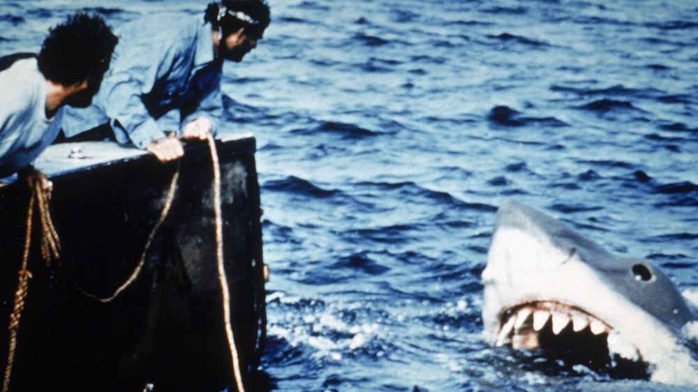 Getty Images / Universal Pictures Deux hommes sur un bateau regardent un requin géant émerger de la mer.