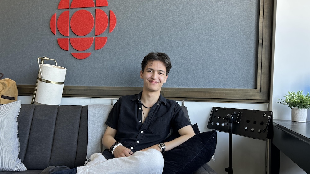 Un homme souriant est assis sur un sofa avec derrière lui, le logo de Radio-Canada.