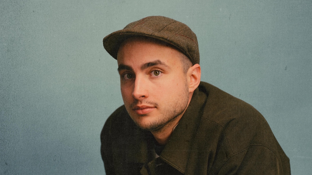 Un homme assis qui porte une casquette classique et une veste olive.