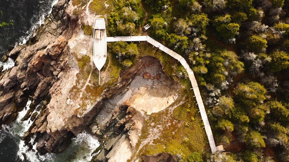 Vue en plongée sur des falaises rocheuses près de l'eau, une structure en bois rappelant un bateau et la forêt.